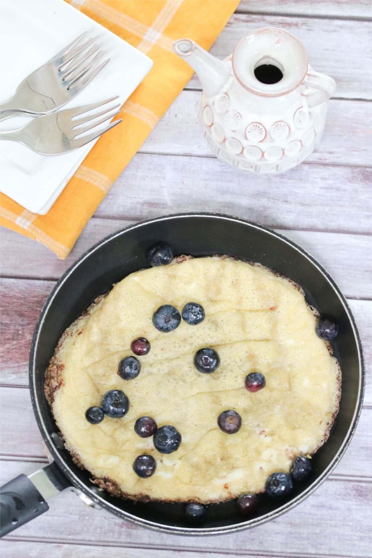 Egg pancake in a black pan with blueberries on top and cutlery behind it.
