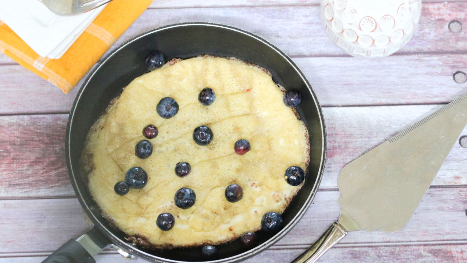 egg pancake in a black pan with blueberries on top and cutlery behind it