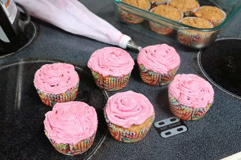 Pink frosted dairy-free smash cupcakes with a piping bag beside them and more cupcakes in a glass tray behind them