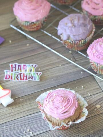 Pink and urple cupcakes with shimmer sprinkles on a cooling rack with a candle number one and happy birthday