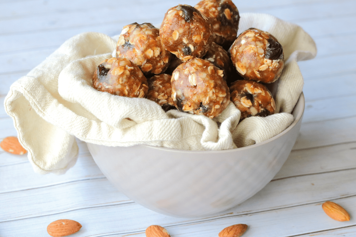 Cherry and almond energy balls in a tan bowl with a linen in it and almonds around