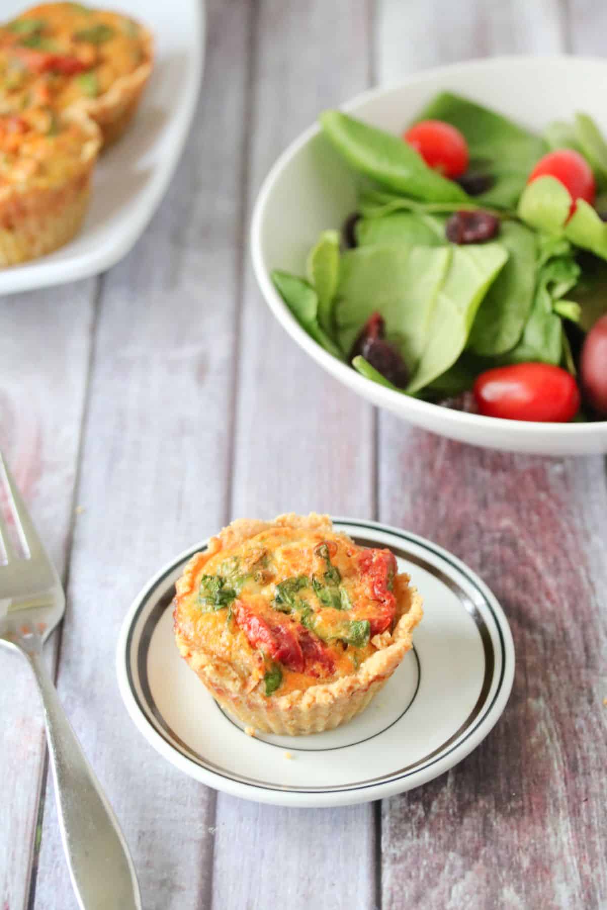 Spinach and mushroom quiche on a white plate with a form, more quiches stacked behind it and a bowl of salad.