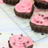 Heart shaped chocolate cupcakes topped with pink icing and chocolate chips on a cooling rack with one cupcake in front.