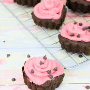Heart shaped chocolate cupcakes topped with pink icing and chocolate chips on a cooling rack with one cupcake in front.