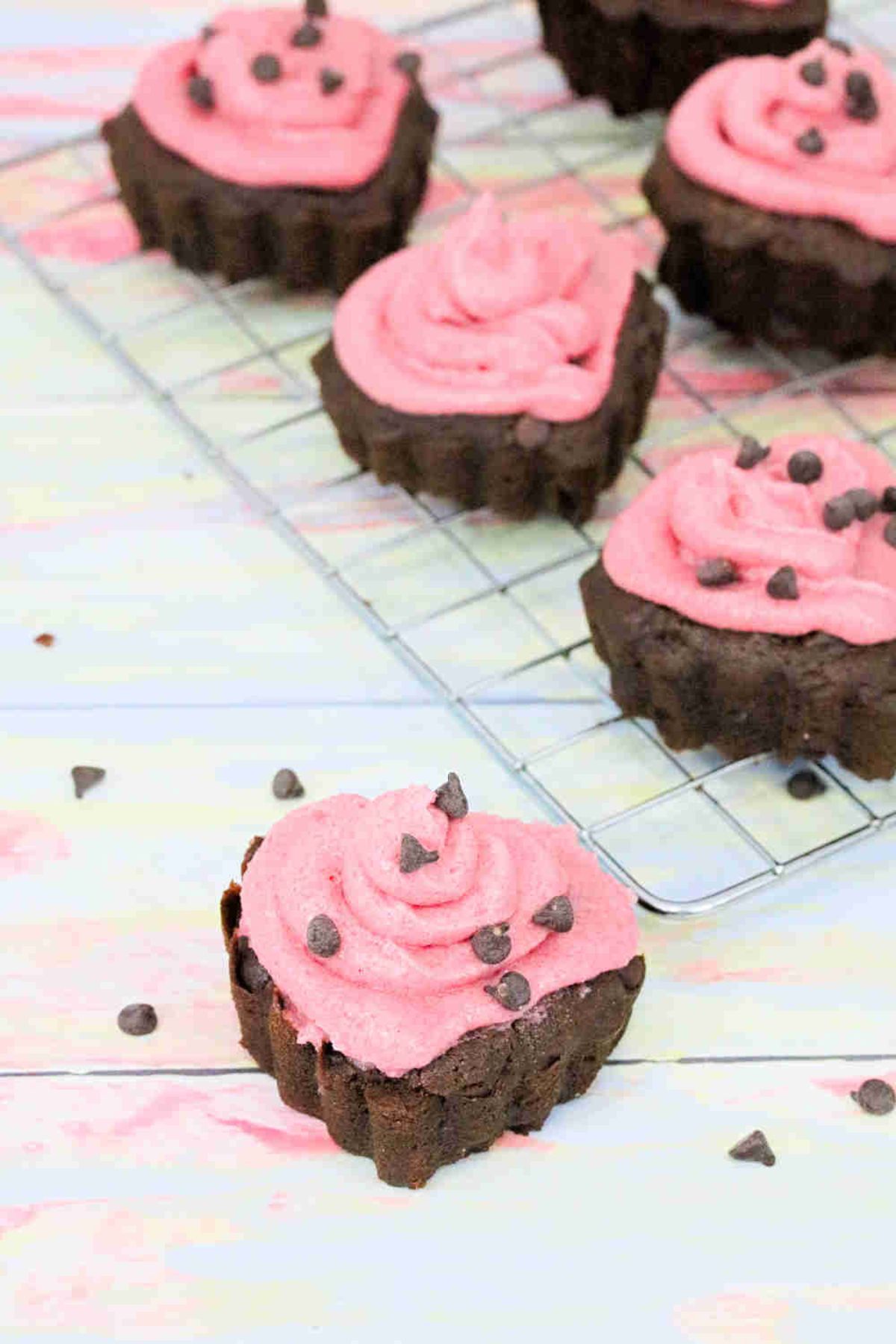 Heart shaped chocolate cupcakes topped with pink icing and chocolate chips on a cooling rack with one cupcake in front.