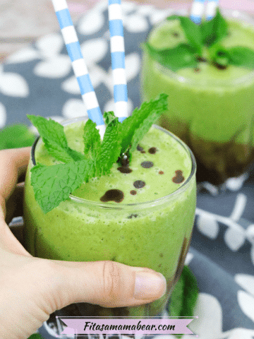 Green mint smoothie topped with fresh mint and chocolate chips with two blue straws and a hand holding the glass