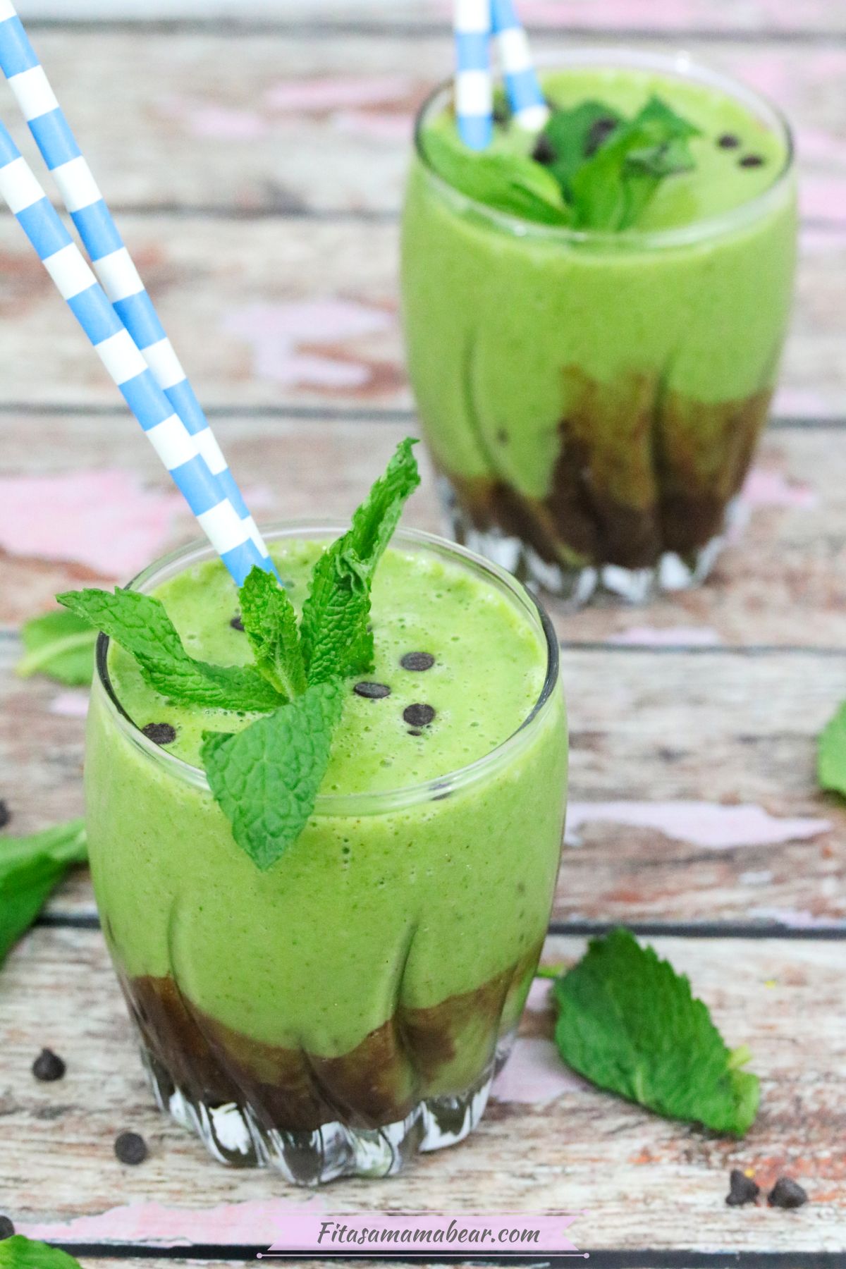 Two glasses with mint green smoothie topped with chocolate chips, fresh mint and blue straws with fresh mint around the glasses.