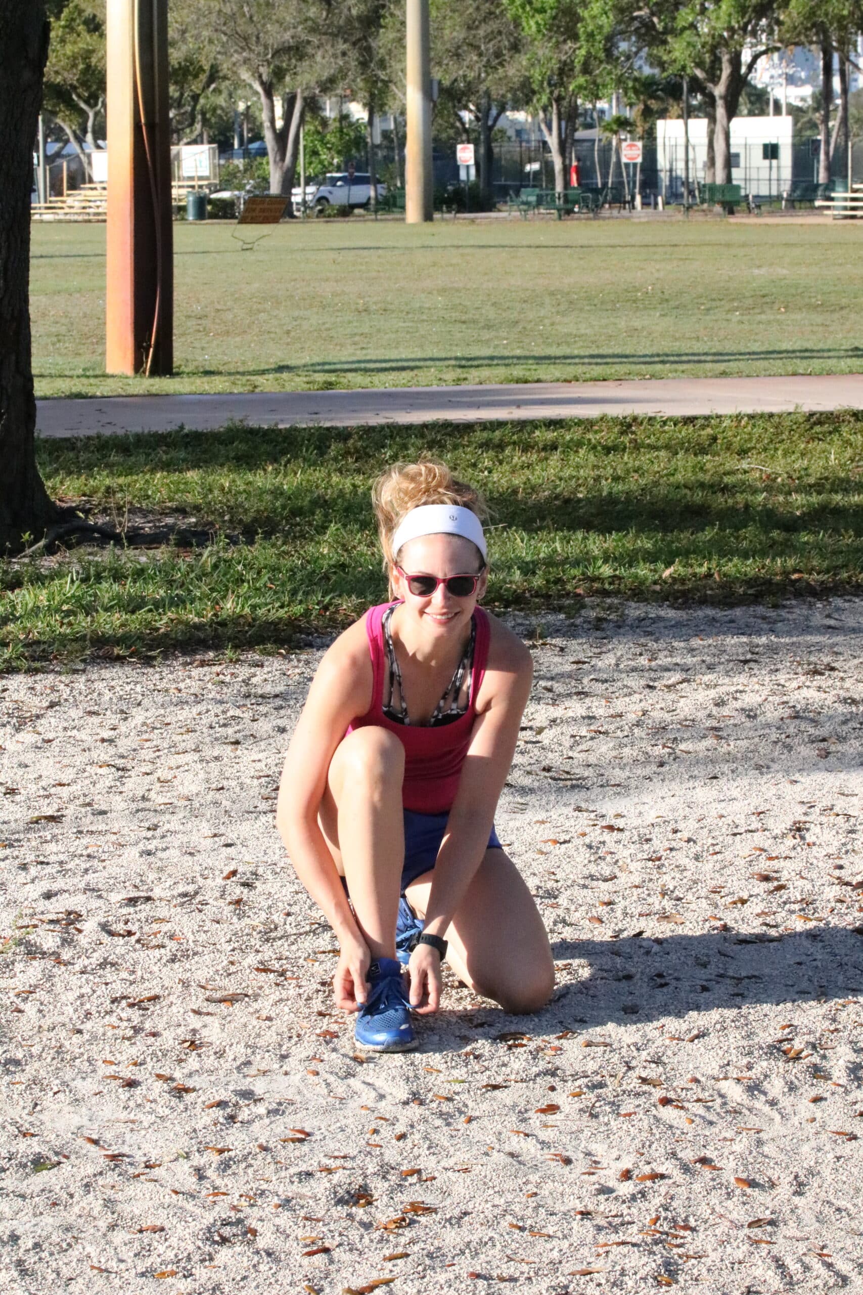 Woman in blue shorts and a maroon shirt with a white headband bending down to tie her shoe in the sand