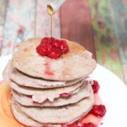 Vegan buckwheat pancakes stacked on a white plate with raspberries on top and syrup being poured on them.