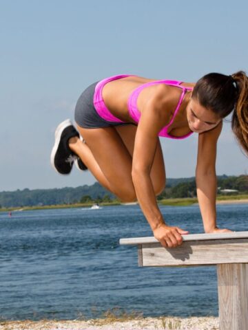 Woman in pink shirt and shorts jumping over a bench on the beach.