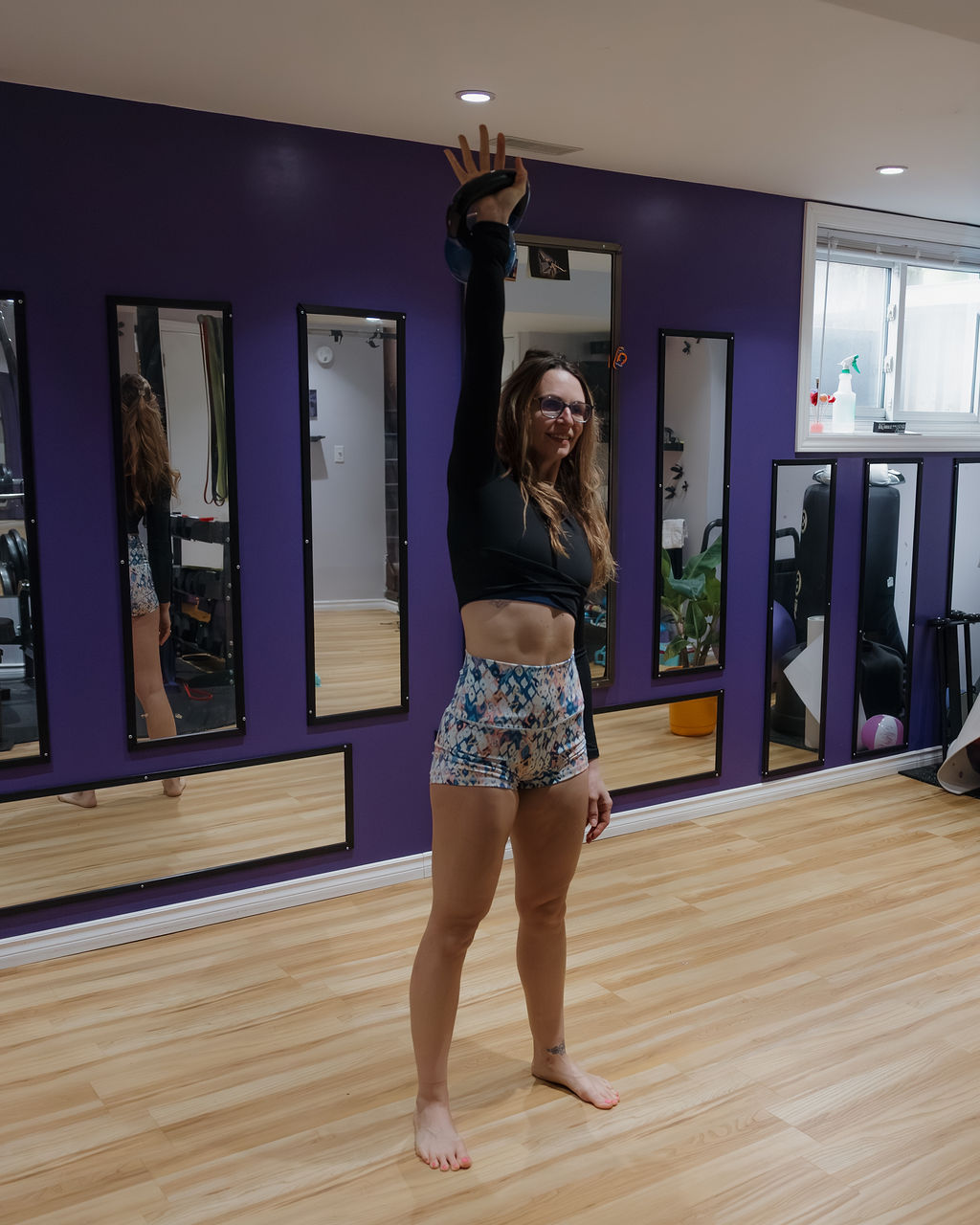 Woman in shorts and a long sleeve crop top performing an overhead kettlebell press.