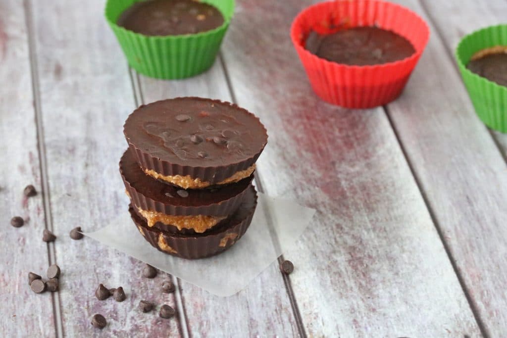 Almond butter chocolate cups stacked in three with more behind it and chocolate chips spread around them