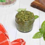 Dairy-free pesto sauce in a mason jar with basil leaves and a plant around it.