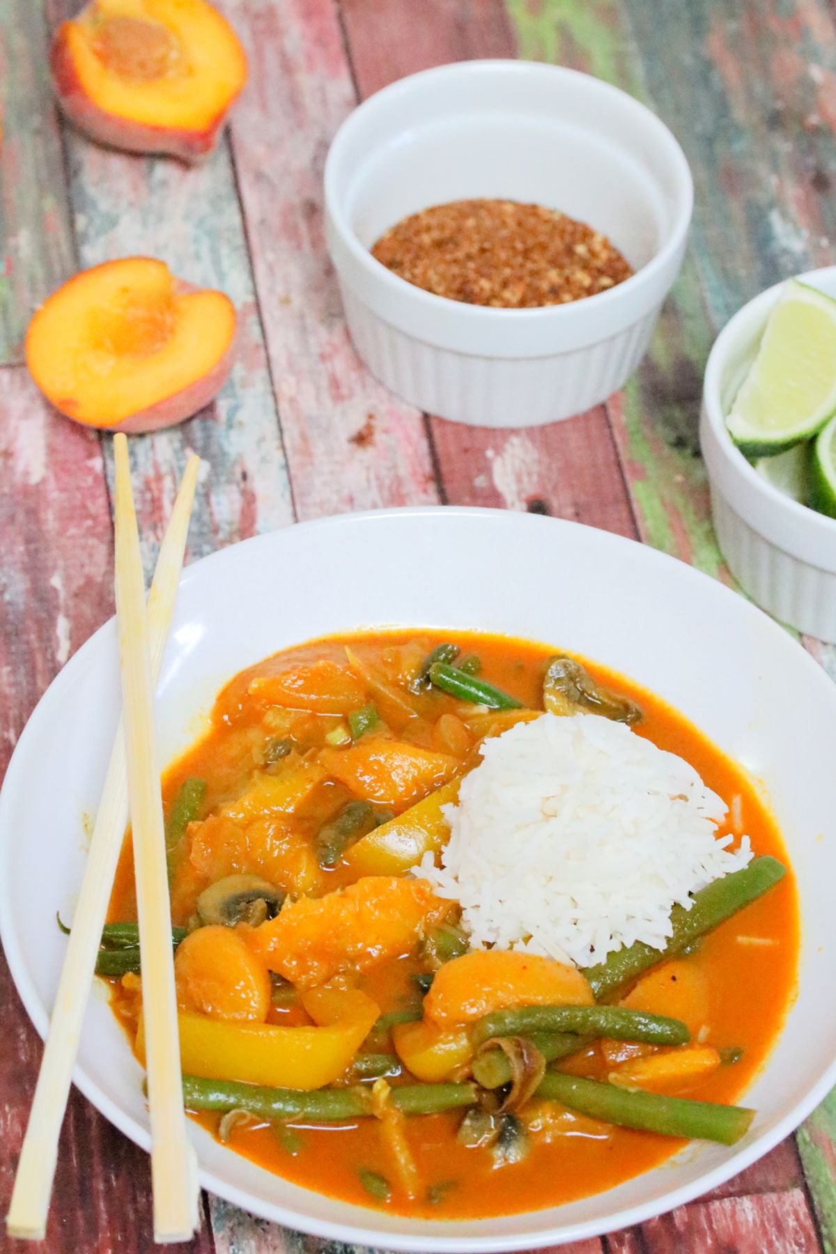 Peach curry in sauce with green beans in a white bowl with rice and seasoning behind it.