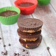 Three chocolate almond butter cups stacked with more butter cups in red and green silicone muffin cups behind them and chocolate chips around them