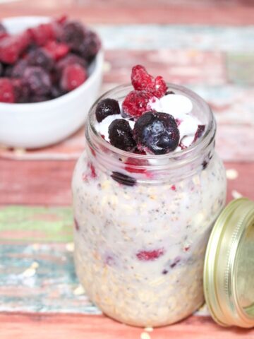 Easy overnight oats in a mason jar topped with frozen berries and coconut whip cream with the jar lid beside it and a bowl of berries behind it.