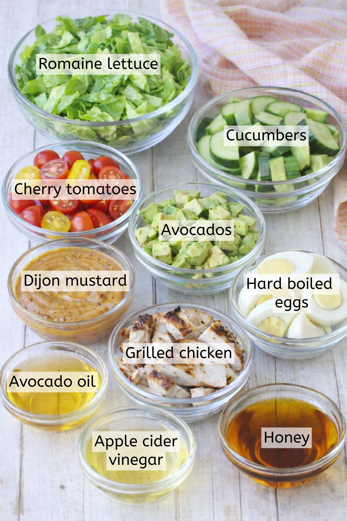 Multiple ingredients to make a cobb salad in glass bowls with labels over top.