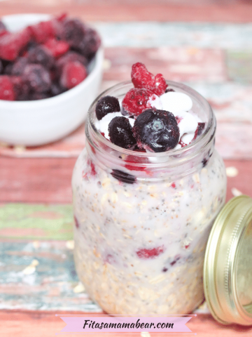 Easy overnight oats in a mason jar topped with frozen berries and coconut whip cream with the jar lid beside it and a bowl of berries behind it