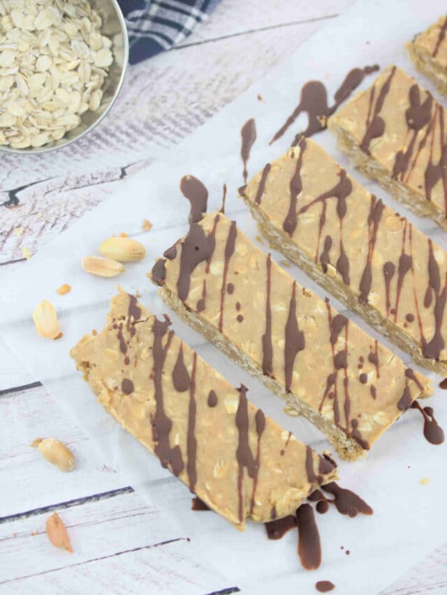 Five peanut butter vanilla protein bars lined up flat on parchment paper and drizzled with chocolate with peanuts and a bowl of oats behind them with a blue linen