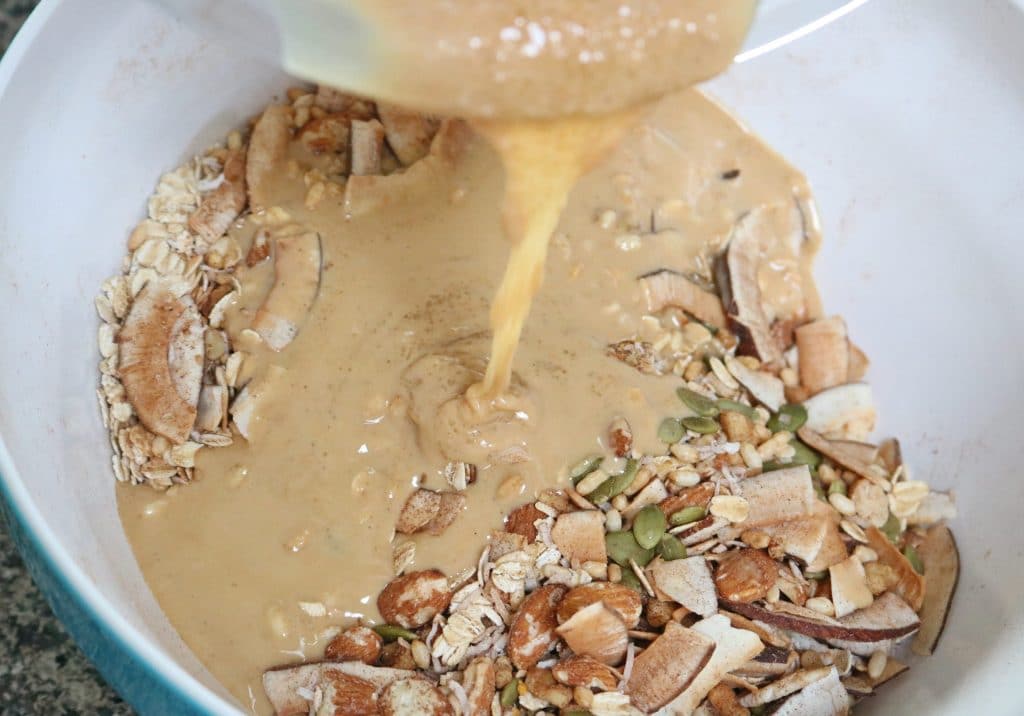 Nuts and seeds in a mixing bowl with cashew butter and coconut oil being poured on top