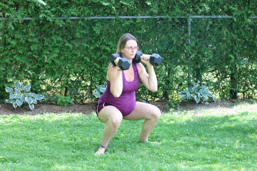 Pregnant woman in pink shirt and black shorts performing a dumbbell squat outside