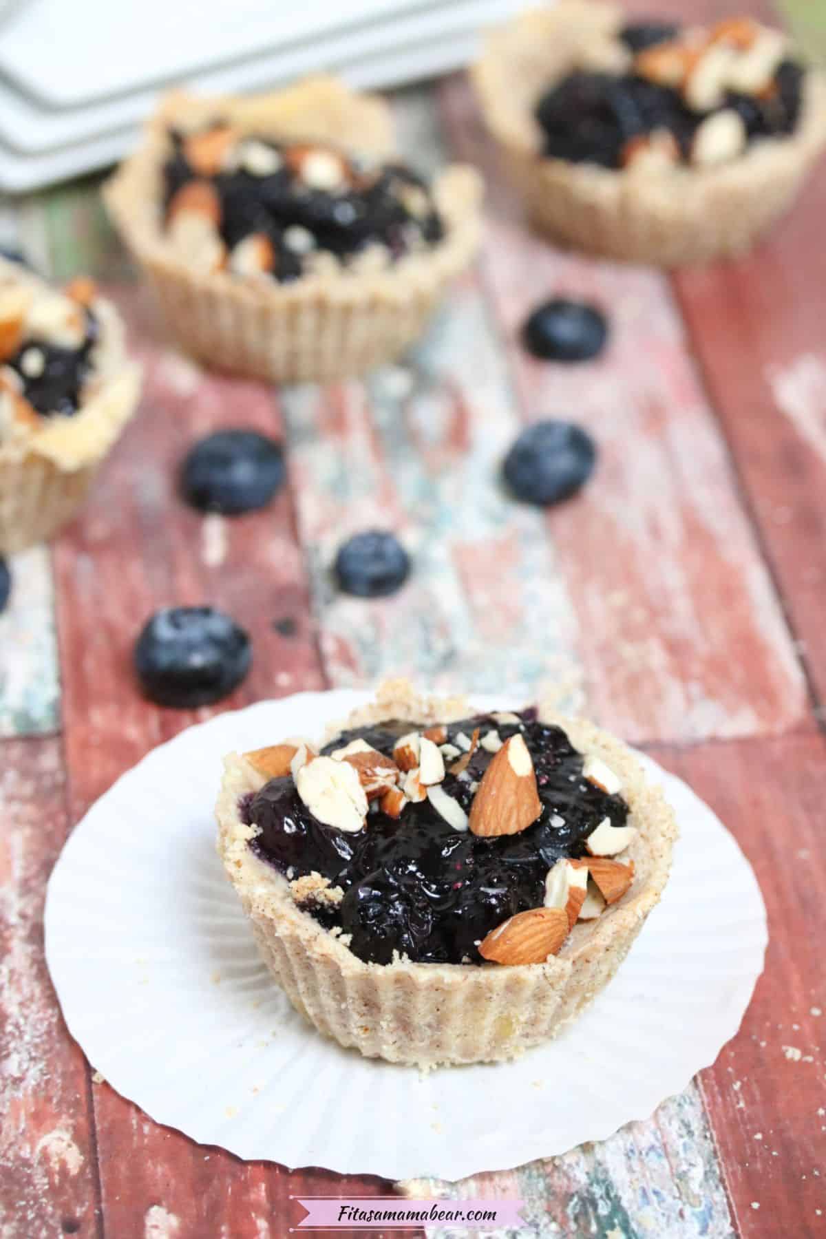 blueberry tart on a white muffin cup with blueberries, more tarts and plates behind it