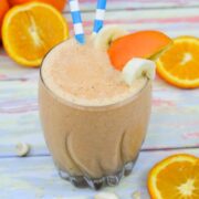 Orange and carrot smoothie in a glass with banana and orange slice on the slide and blue straws in it.