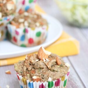 almond zucchini muffin in a polkadot muffin cup in front of more muffins on a white plate and yellow napkin