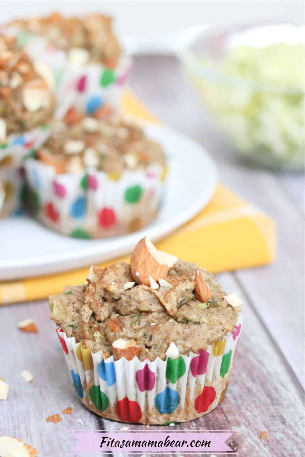 almond zucchini muffin in a polkadot muffin cup in front of more muffins on a white plate and yellow napkin