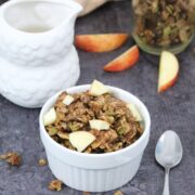 Apple pie granola in a white ramekin with a spoon beside it, a milk jug, more granola and fresh sliced apple behind it.