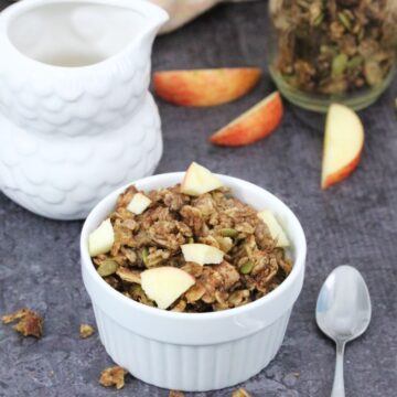 Apple pie granola in a white ramekin with a spoon beside it, a milk jug, more granola and fresh sliced apple behind it.