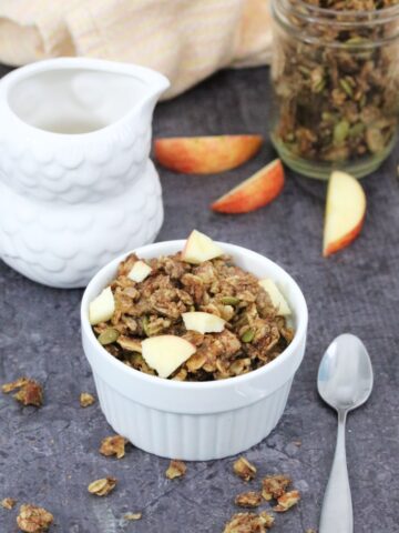 Apple pie granola in a white ramekin with a spoon beside it, a milk jug, more granola and fresh sliced apple behind it.