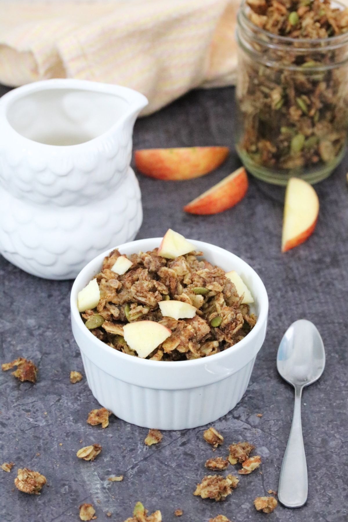 Apple pie granola in a white ramekin with a spoon beside it, a milk jug, more granola and fresh sliced apple behind it.