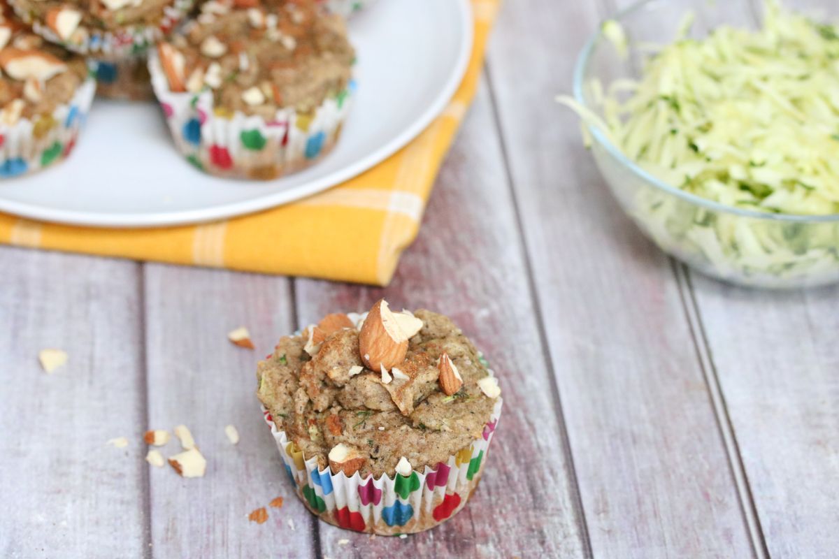 almond zucchini muffin in a polkadot muffin cup in front of more muffins on a white plate and yellow napkin