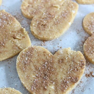 Coconut flour sugar cookies in heart shapes on parchment paper sprinkled with cinnamon and sugar.