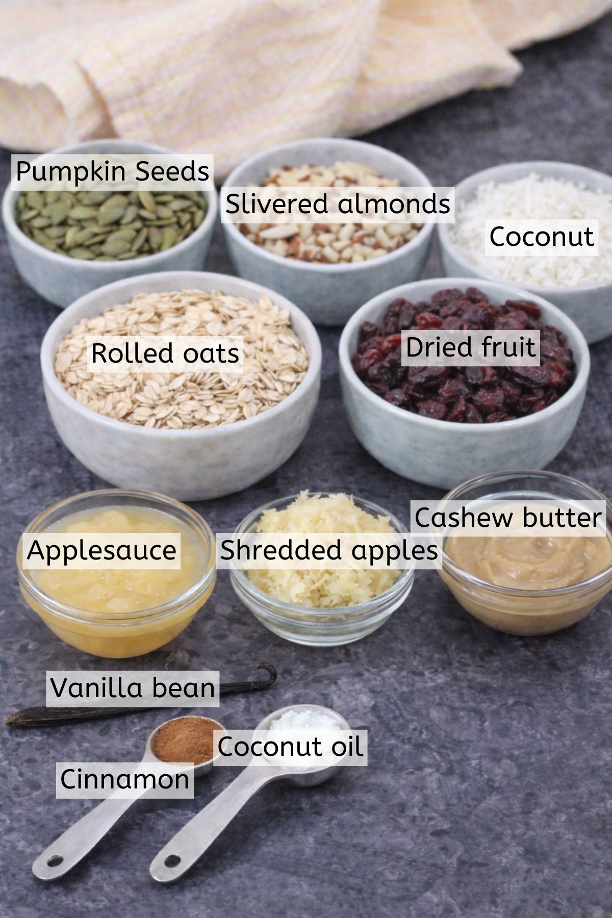 Granola ingredients like oats, coconut, nuts, and seeds in bowls with text labels over top.
