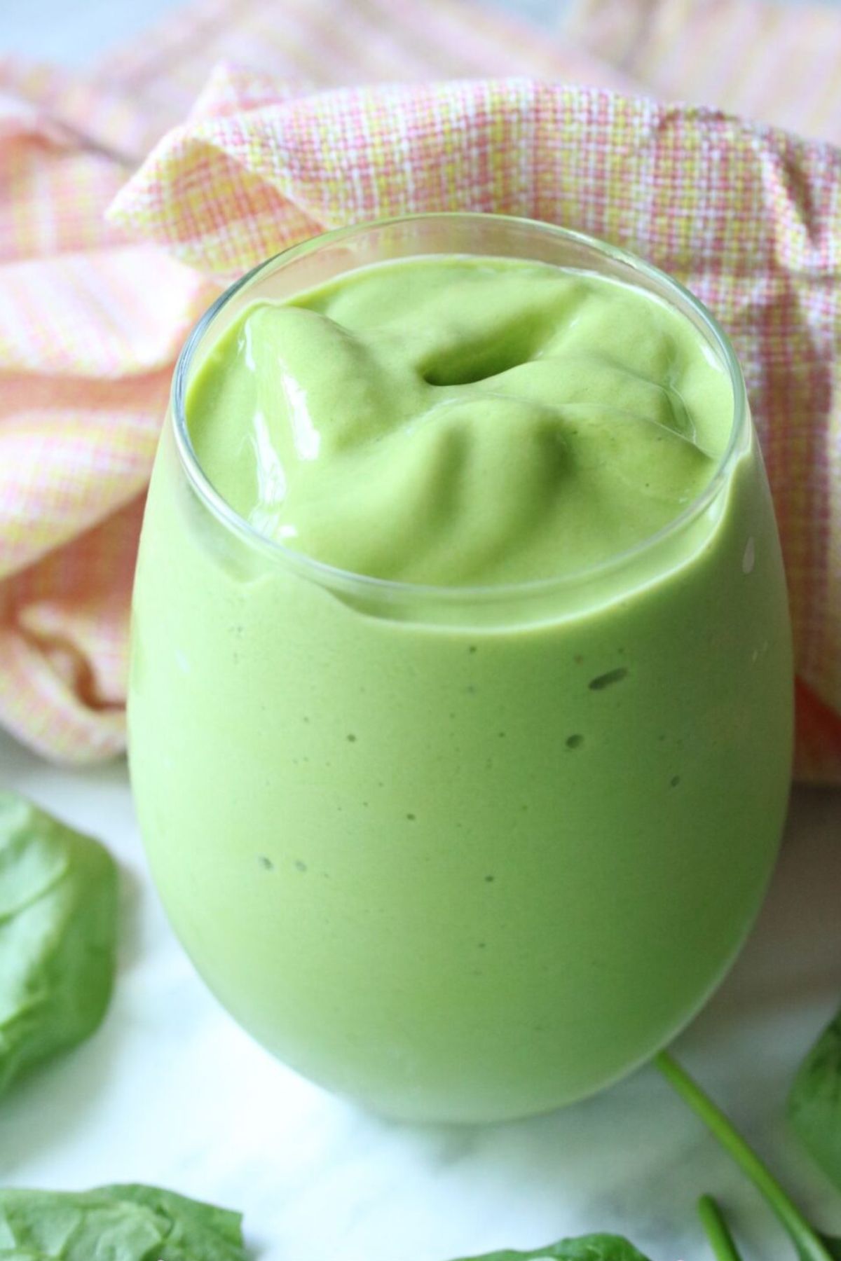 Mango green smoothie in a tumbler glass with spinach leaves around it and an orange linen behind it.