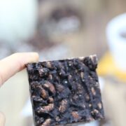Two fingers holding up a square of a dark chocolate and peanut butter crunch bar.