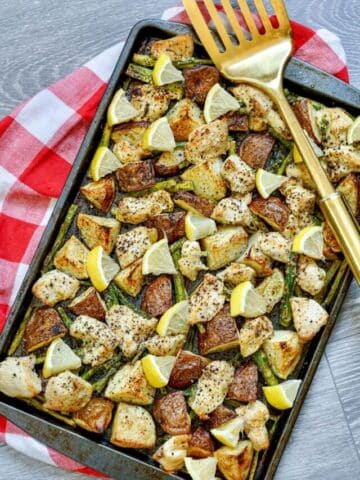 Roasted chicken, potatoes, and asparagus with lemons on a baking tray on a checkered red linen.