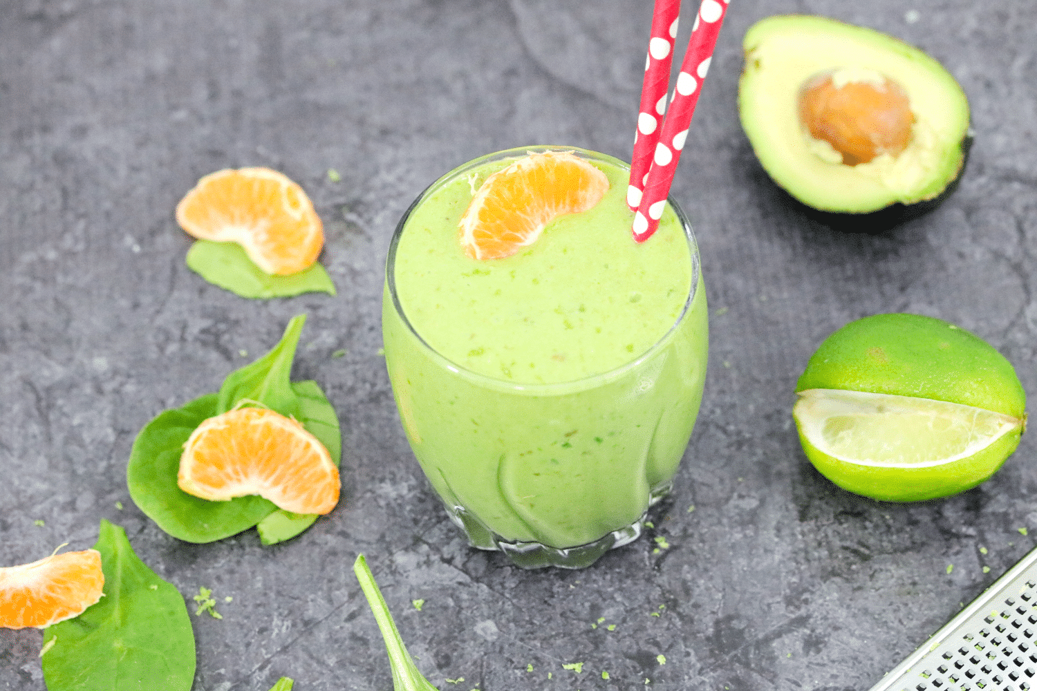 tropical green smoothie with red straws in the glass and orange, spinach, avocado and lime around it