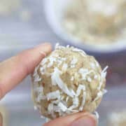 Two fingers holding a coconut protein ball with a purple bowl of almonds and coconut behind it