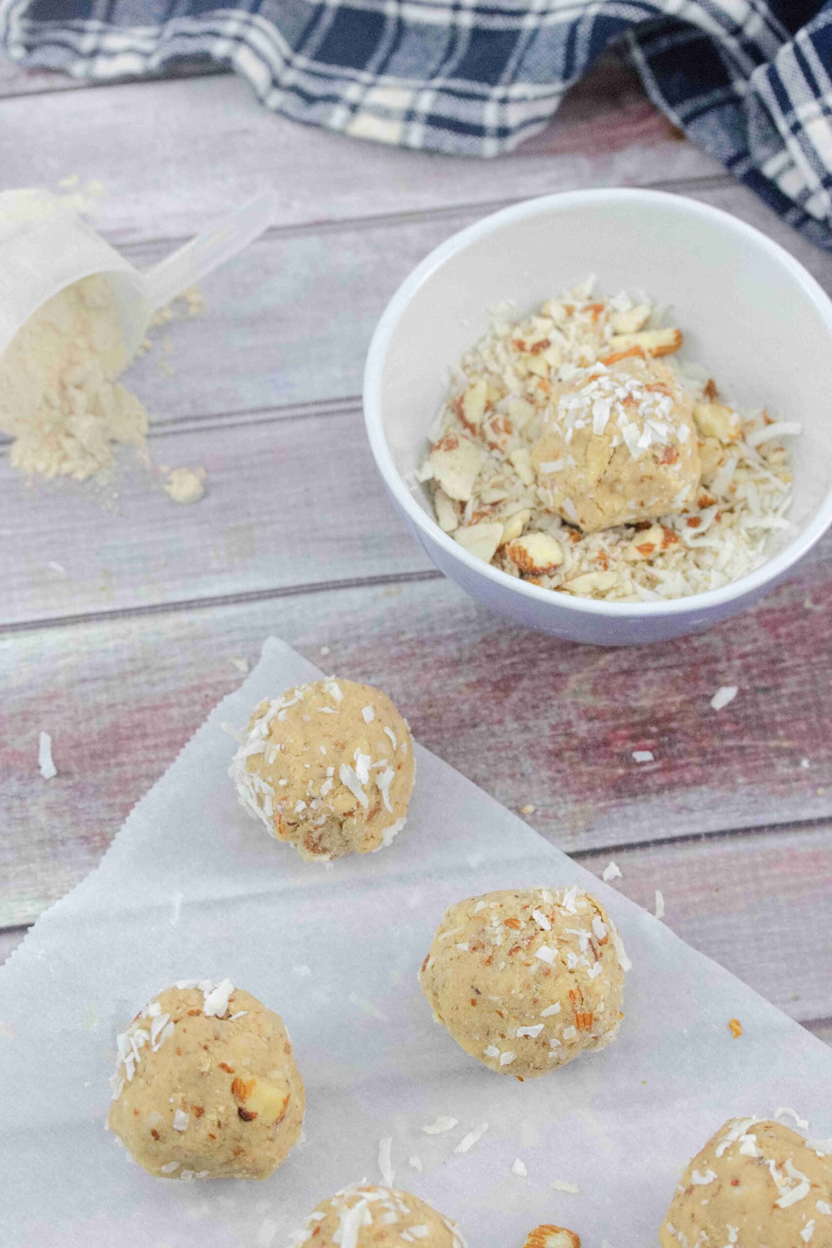 Multiple gluten-free protein balls rolled in coconut on a piece of white parchment paper with a bowl of coconut and almonds and a scoop of protein powder behind them