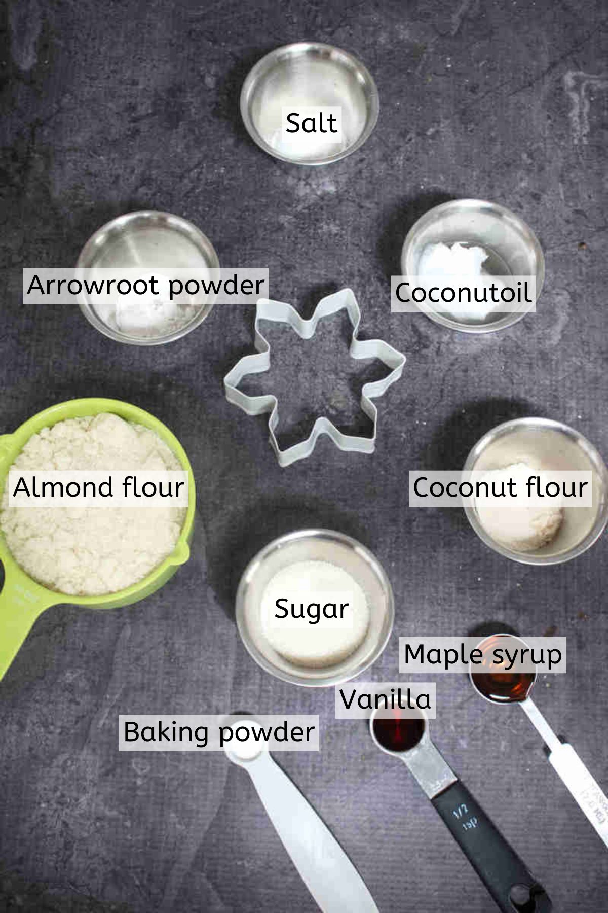 Cookie Ingredients like almond flour, sugar, maple syrup and vanilla in measuring cups with text labels over top.