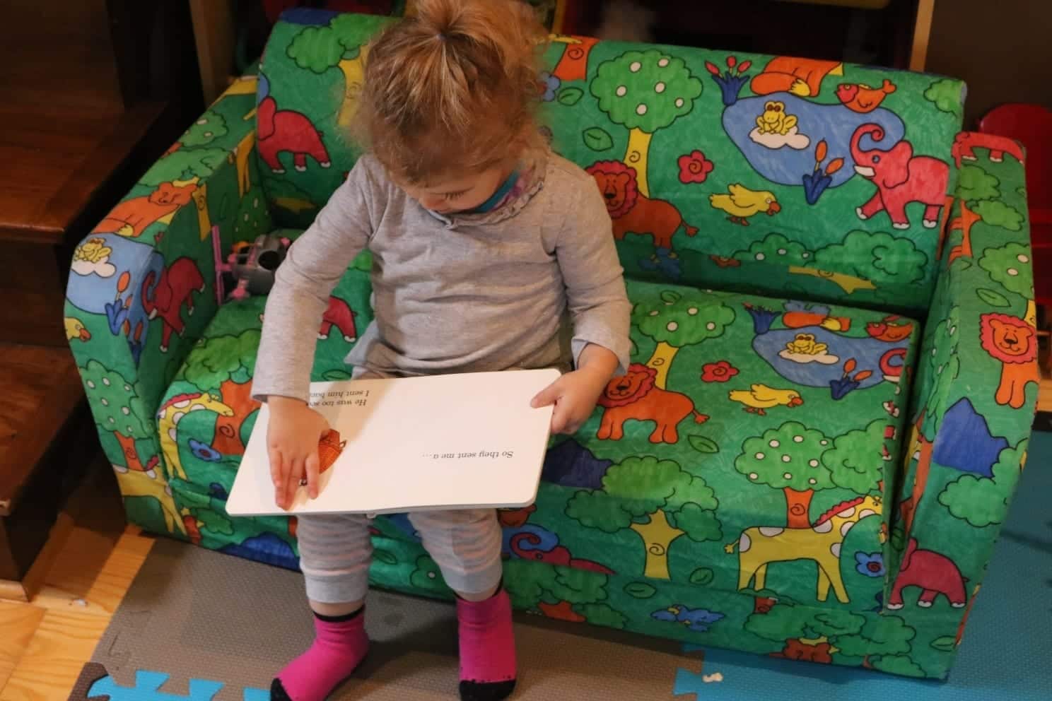 Toddler sitting on a toddler couch in a grey outfit reading a lift the flap book