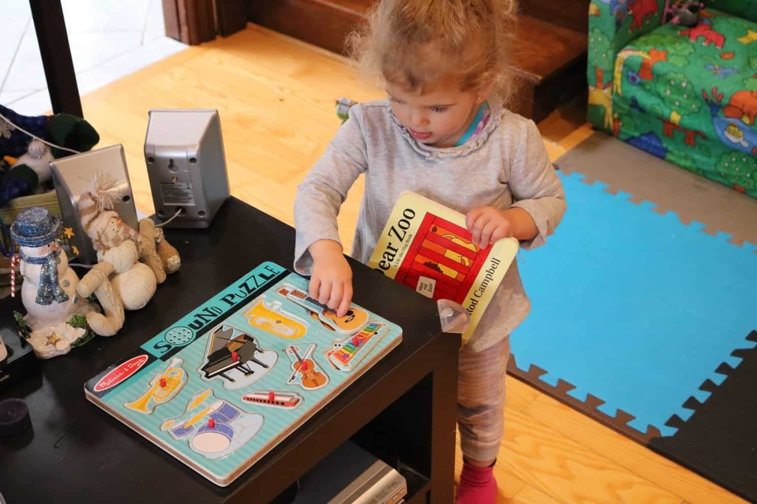 Toddler in a grey shirt playing with a noise puzzle while holding a book