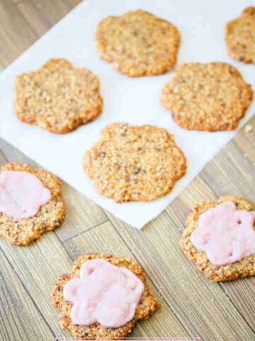 Multiple crispy sugar cookies topped with pink icing and cane sugar with some of the cookies on parchment paper.