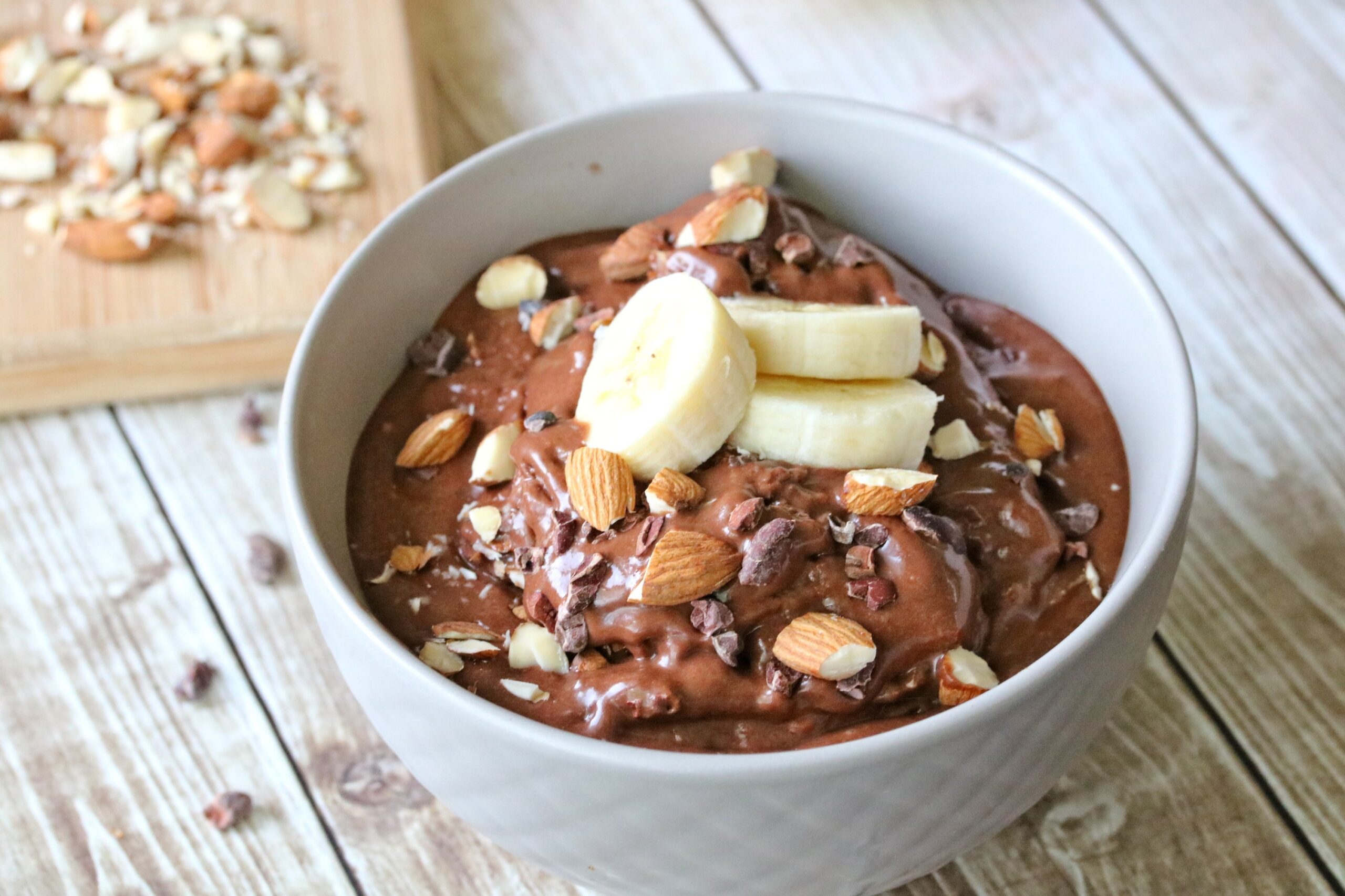 Grey bowl with chocolate banana nice cream topped with sliced bananas and almonds with more chopped almonds on a cutting board behind it