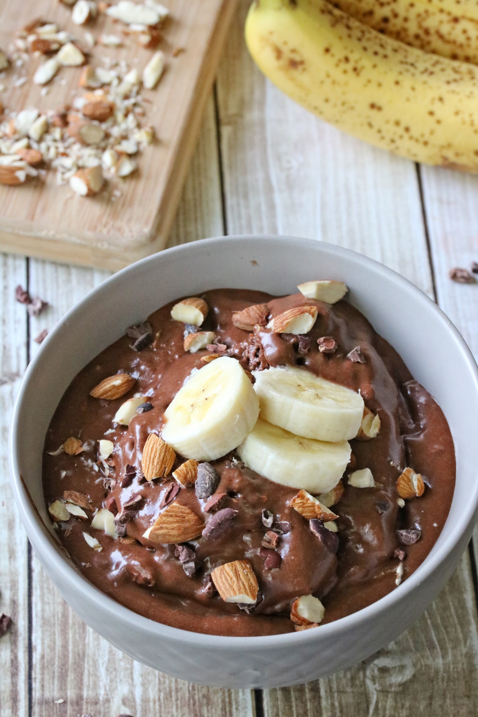 Grey bowl with chocolate banana nice cream topped with sliced bananas and almonds with more chopped almonds on a cutting board behind it