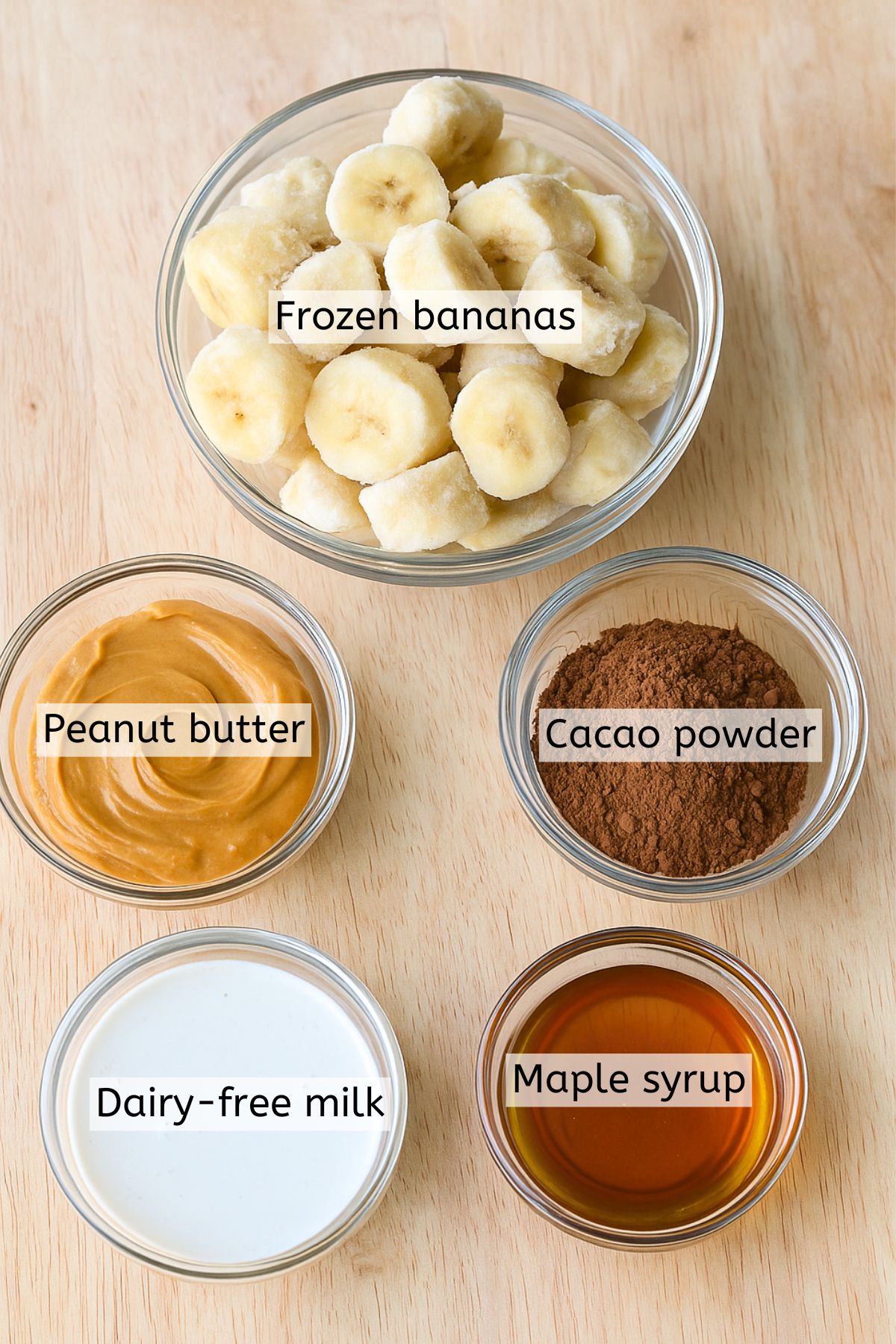Frozen bananas, milk, cacao powder, and maple syrup in glass dishes with text labels over top.