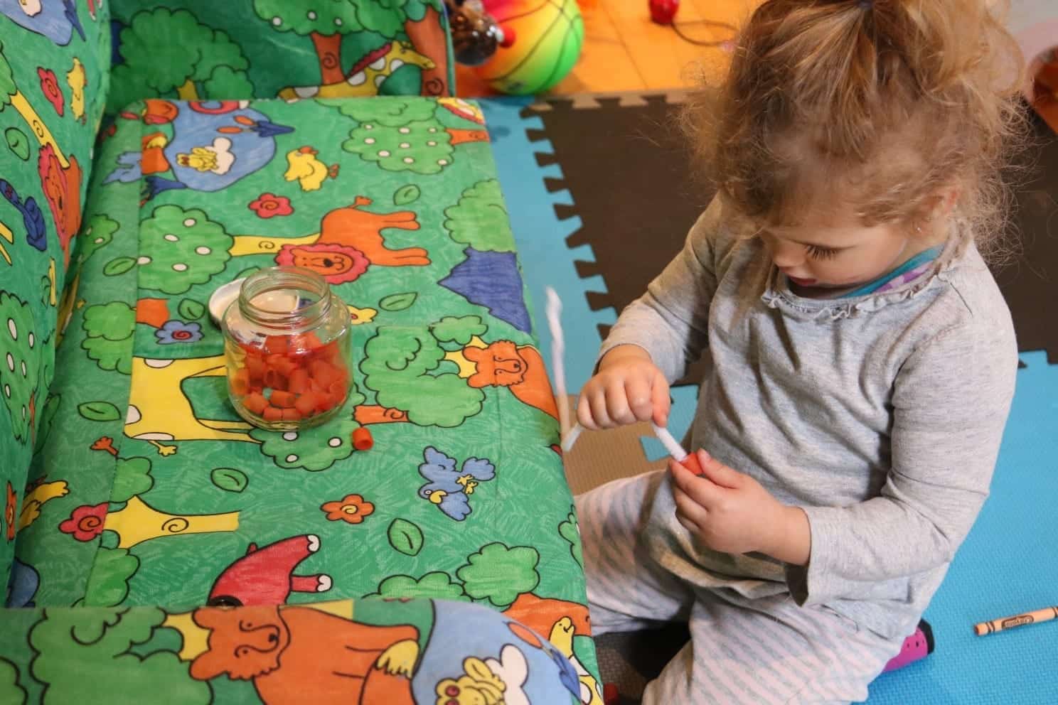 Toddler in grey shirt and pants threading colored noodles onto a pipe cleaner